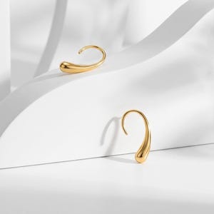 Könnte beinhalten: Ein Paar goldfarbene Ohrringe in einer glatten, polierten Tropfenform. Jeder Ohrring hat ein geschwungenes Design mit offener Schlaufe, das spitz zuläuft und eine minimalistische Silhouette mit einer reflektierenden Oberfläche bildet.