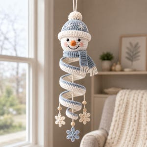 Könnte beinhalten: Ein gehäkelter Schneemann als Hängedekoration mit einem blau-weißen spiralförmigen Körper, einer passenden blauen Mütze mit weißem Bommel und Schneeflockenstickerei, einem blauen Schal und drei hängenden, an Holzperlen befestigten Häkelschneeflocken.
