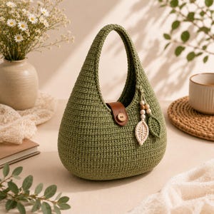 Puede incluir: Un bolso de ganchillo en color verde salvia con forma de gota y asa integrada. El bolso cuenta con una lengüeta de cierre de aspecto cuero marrón asegurada con un botón de madera y un adorno colgante de cuentas de madera y dos hojas tejidas.
