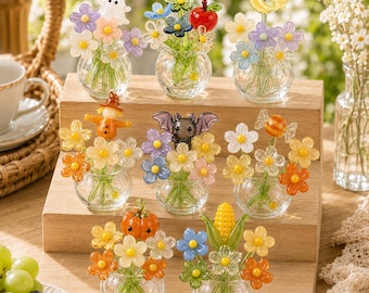Puede incluir: Una colección de ocho jarrones de vidrio en miniatura, cada uno con un pequeño ramo de flores de vidrio de colores y una figura estacional única, incluyendo un fantasma, manzana, luna, espantapájaros, murciélago, caramelo, calabaza y maíz, dispuestos sobre un soporte de madera.