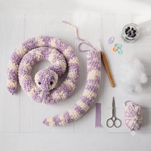 May include: A coiled plush snake made of purple and cream variegated yarn with black safety eyes and a purple felt tongue. The set includes a crochet hook, a small jar of safety eyes, stitch markers, scissors, fibre fill, and yarn.