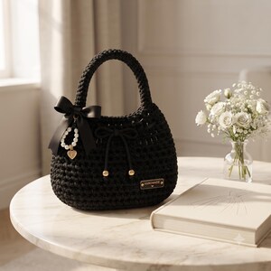 May include: A black crocheted handbag with a rounded handle, featuring a black satin bow, a pearl-accented charm with a heart pendant, a drawstring closure with gold-toned beads, and a small rectangular tag labeled HANDMADE in gold lettering.