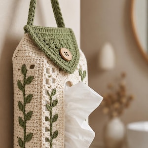 May include: A cream-colored crocheted tissue box cover featuring a green leaf vine pattern on the sides, a triangular flap with a wooden button closure, and a matching green crocheted hanging strap, holding a white tissue.