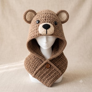 Pode incluir: Um capuz de crochê castanho em forma de urso com focinho creme, olhos de botão pretos e nariz bordado a preto. O capuz apresenta orelhas arredondadas e um fecho de botão castanho na frente, com um padrão de pontos texturizado.