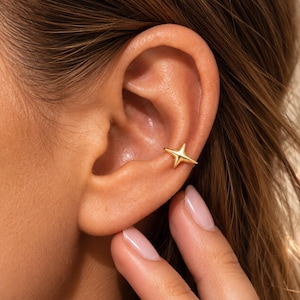 May include: A gold-toned ear cuff shaped like a four-pointed star. The jewelry piece features a smooth, polished surface and is positioned securely on the concha area of the ear, showcasing a simple, symmetrical design with sharp, tapered points.