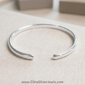 May include: A polished silver cuff bracelet with a smooth, rounded surface and an open design. The bracelet features a simple, continuous band that curves into a circular shape. The text www.ElinaSilverJewls.com is printed at the bottom.