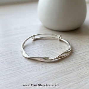 May include: A silver bangle bracelet with a smooth, undulating, wavy surface texture. The metal band features a continuous, flowing design that curves gently around its circumference. The text www.ElinaSilverJewls.com is printed at the bottom.
