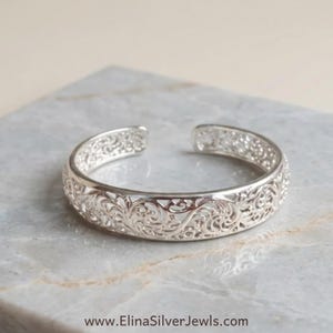 Peut inclure: Un bracelet manchette en argent présentant un motif filigrane ajouré complexe avec des volutes. Le bracelet possède une finition métallique polie et une ouverture à l'arrière. Le texte www.ElinaSilverJewels.com est visible.