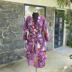May include: A long-sleeved robe featuring a patchwork design in various shades of purple, accented with floral and geometric patterns in white, orange, and blue. The garment includes a matching tie belt at the waist and two front patch pockets.