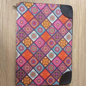May include: A rectangular zippered portfolio case featuring a vibrant, multi-colored diamond pattern with intricate circular mandala-style motifs in shades of orange, pink, teal, and purple, accented by solid black leather-like corner reinforcements.