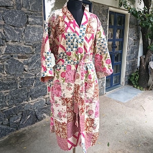 May include: A long-sleeved quilted robe featuring a patchwork design with various floral and geometric patterns in shades of pink, brown, and cream. The garment includes a matching fabric tie belt at the waist and a V-neckline, displayed on a mannequin.