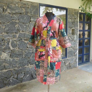 Puede incluir: Una bata acolchada hasta la rodilla con un diseño de patchwork compuesto por varios cuadrados de tela en tonos rojo, amarillo, rosa, verde y negro. La prenda incluye mangas largas, un cinturón a juego en la cintura y dos bolsillos frontales.