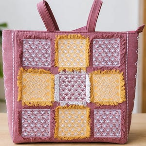 Può includere: Una borsa tote color malva con un design patchwork di toppe di tessuto quadrate dai bordi sfilacciati. Le toppe si alternano tra motivi a ventaglio ricamati in giallo senape e bianco. La borsa ha due manici e una chiusura a cerniera.