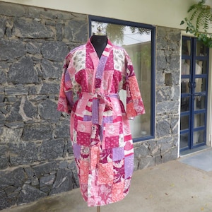 Könnte beinhalten: Ein knielanger Kimono-Bademantel mit einem Patchwork-Design aus verschiedenen Blumen- und geometrischen Mustern in Rosa-, Magenta- und Weißtönen. Das Kleidungsstück hat lange Ärmel, einen V-Ausschnitt und einen passenden Stoffgürtel.