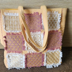 Può includere: Una borsa tote color arancione chiaro caratterizzata da una griglia di nove toppe quadrate con bordi sfrangiati. Le toppe alternano ricami dorati e color malva con vari motivi geometrici. La borsa ha due manici color arancione chiaro.