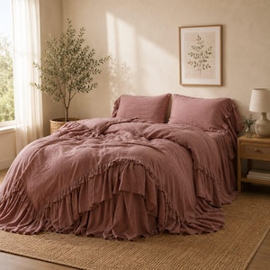 May include: A dusty rose bedding set featuring a duvet cover with ruffled edges and two matching pillow shams. The fabric has a textured appearance, and the set is draped over a bed, creating a layered look with soft, cascading fabric ruffles.