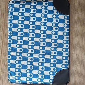 May include: A rectangular laptop sleeve featuring a repeating blue and white geometric pattern of interlocking rounded shapes. The sleeve includes dark brown faux leather corner accents on the top right and bottom right sides with a zipper closure.