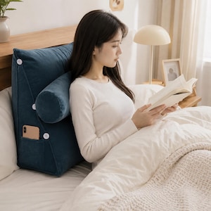 May include: A dark teal, wedge-shaped velvet reading pillow featuring a detachable cylindrical bolster, three white decorative buttons along the side, and a small side pocket containing a smartphone. The pillow is designed for back support.