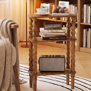 May include: A three-tiered wooden side table with four turned, bobbin-style legs. The table features a warm brown wood grain finish, three rectangular shelves, and a sturdy frame. It is currently holding a white bowl, a small tray, books, a digital clock, and a rectangular speaker.