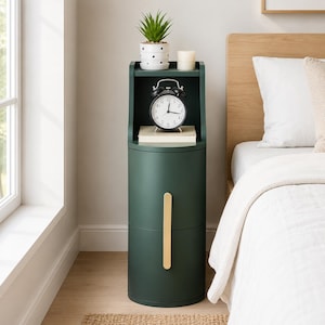 May include: A tall, cylindrical dark green bedside table featuring a vertical gold handle on the front door. The top shelf holds a small potted plant in a white polka-dot pot, a white pillar candle, a black alarm clock, and a stack of books.