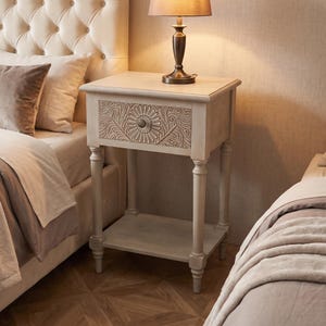 May include: A light-colored wooden nightstand featuring a single drawer with an intricate floral carved pattern and a central knob. The piece stands on four turned legs and includes a lower open shelf for storage, topped with a metal table lamp.