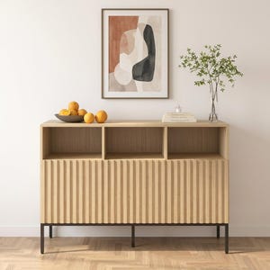 May include: A light wood sideboard featuring three open upper compartments and three lower cabinet doors with vertical slats. The cabinet is supported by a thin black metal frame. A bowl of oranges and a glass vase with green branches sit on top.