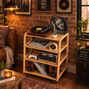 May include: A light-colored wooden four-tier rolling storage cart featuring a turntable on the top shelf, vinyl records, a small speaker, a game controller, a larger audio speaker, and a small potted cactus on the shelves.