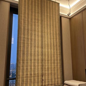 May include: A rectangular window shade featuring a woven tan and dark green herringbone pattern with a solid dark green fabric border. The shade is suspended from a ceiling mount and includes a thin vertical cord for adjustment and operation.