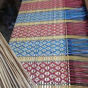 May include: A hand-woven mat on a loom featuring a repeating geometric pattern in red, blue, and yellow threads interlaced with light-colored reeds. The mat displays vibrant horizontal stripes and loose, colorful warp threads extending from the edge.