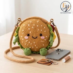 May include: A round, crocheted hamburger-shaped crossbody bag with a tan bun featuring white embroidered sesame seeds, green lettuce, and a smiling face with black eyes and pink cheeks. It includes a matching tan crocheted shoulder strap.