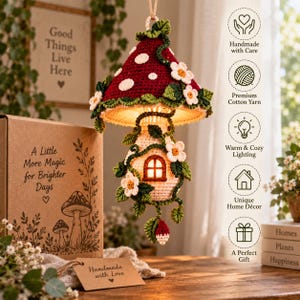May include: A hanging crochet mushroom house lamp with a dark red cap featuring white spots, a cream-colored stem, and green vines with white flowers. The lamp emits a warm glow from a small window. It is presented with a cardboard gift box.