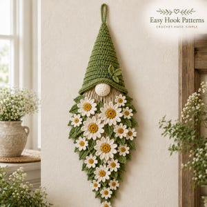 Op de afbeelding: Een wandhanger met een gehaakte kabouter met een hoge groene muts, een ronde beige neus en een lange baard versierd met talloze witte en gele madeliefjes en groene gehaakte bladeren. De tekst Easy Hook Patterns is zichtbaar.
