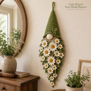 Op de afbeelding: Een gehaakte wandhanger in de vorm van een kabouter met een hoge groene puntmuts met een klein blaadje, een ronde beige neus en een lange baard bestaande uit talloze witte en gele madeliefjes afgewisseld met groene gehaakte bladeren. Tekst: Easy Hook Patterns.