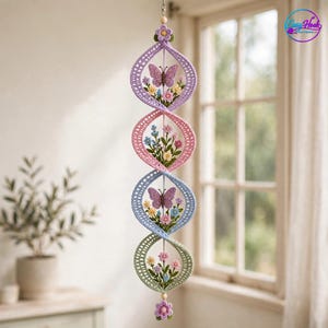Puede incluir: Un móvil colgante vertical con cuatro segmentos en espiral de ganchillo interconectados en tonos lavanda, rosa, azul y verde salvia. Cada segmento contiene motivos florales bordados y mariposas moradas, rematados con pequeñas flores de ganchillo.