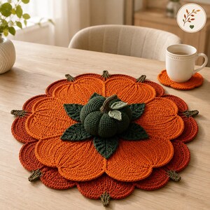 May include: A round, orange, crocheted pumpkin-shaped placemat featuring a dark green, three-dimensional crocheted pumpkin in the centre with green leaves. A matching small, orange, scalloped crocheted coaster sits under a white ceramic mug.