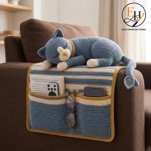 Könnte beinhalten: Ein gehäkeltes blaues Katzen-Plüschtier auf einem gestreiften Armlehnen-Organizer in Blau und Creme. Der Organizer hat zwei Taschen, die ein Buch, eine Fernbedienung, ein Smartphone und eine Brille enthalten. Die Katze hat cremefarbene Pfoten und Schnauze.