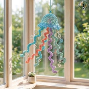 May include: A crocheted jellyfish hanging decoration featuring a light blue bell-shaped top with intricate lace-like patterns. It has multiple long, spiraling tentacles in shades of teal, coral, and purple, accented with small white crocheted rings.