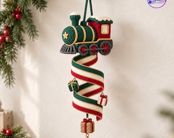 May include: A crocheted Christmas train ornament in dark green, red, and white, featuring a spiral ribbon tail adorned with three small gift boxes and a gold metal bell at the bottom. The train has a yellow star on the front and a white roof.