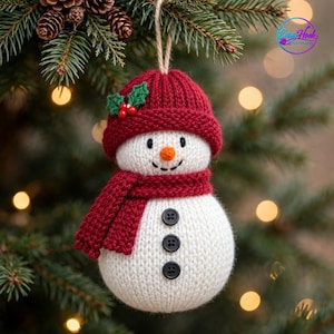 May include: A white knitted snowman ornament featuring a red ribbed beanie with green holly leaves and red berries, a matching red scarf, and three black buttons down the front. The snowman has a small orange nose and black stitched eyes and mouth.