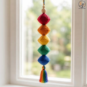 May include: A hanging decoration featuring five stacked, diamond-shaped crocheted ornaments in red, orange, yellow, green, and blue. A smooth, round wooden bead sits at the top, and a multi-colored yarn tassel hangs from the bottom of the structure.