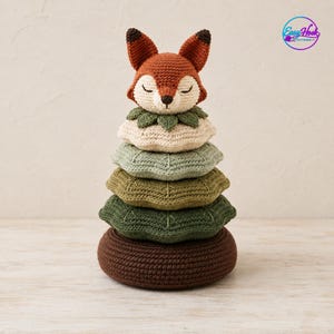 Peut inclure: Un jouet à empiler en crochet composé d'une tête de renard aux yeux fermés et oreilles pointues, reposant sur cinq anneaux empilés à bords festonnés dans des tons crème, vert clair, vert sauge, vert foncé et marron foncé.