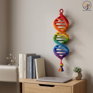 May include: A crocheted DNA double helix wall hanging featuring a vibrant rainbow gradient of red, orange, yellow, green, blue, and purple yarn. The structure consists of two twisted strands connected by horizontal rungs, ending in a small tassel.