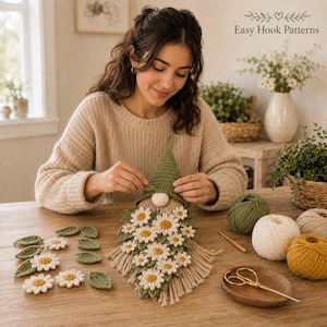 Op de afbeelding: Een handgemaakte gehaakte kabouter wandhanger met een groene puntmuts, een ronde beige neus en een baard versierd met witte en gele madeliefjes en groene blaadjes. Het item heeft een franje onderkant en ligt tussen verschillende bollen garen.