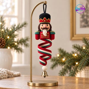 Op de afbeelding: Een gehaakte notenkraker-ornament hangend aan een gouden metalen standaard. De notenkraker heeft een zwarte hoed, een rood jasje met gouden details en een witte baard. Onder de figuur hangt een spiraalvormige rood-wit-groene staart met een belletje.