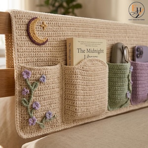 May include: A beige crochet bedside organizer featuring four pockets. The first pocket displays a purple floral vine, the second holds a book titled The Midnight Library, the third contains glasses, and the fourth holds a purple smartphone. A yellow crescent moon is stitched on the top left corner.
