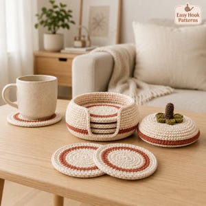 Op de afbeelding: Een set ronde, crèmekleurige gehaakte onderzetters met een roestkleurig cirkelvormig accent. De set bevat een bijpassende ronde houder met een deksel bekroond met een groen blaadje en een bruin steeltje, en een gespikkelde keramische mok.