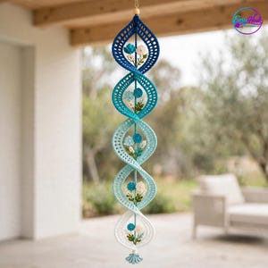 Op de afbeelding: Een verticaal hangend gehaakt spiraalornament met een kleurverloop van donkerblauw aan de bovenkant naar wit aan de onderkant. De structuur bevat kleine gehaakte bloemen in blauw en wit met groene bladeren als accenten.