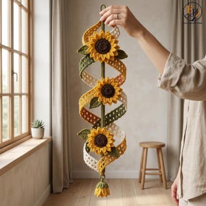 Op de afbeelding: Een verticale gehaakte wandhanger met drie gele zonnebloemen met donkerbruine harten, afgewisseld met groene gehaakte bladeren en een spiraalvormig, opengewerkt lint in tinten crème, geel en olijfgroen, eindigend in een kleine kwast.