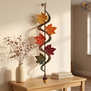 May include: A vertical wall hanging featuring a crocheted spiral vine adorned with five maple leaves in shades of yellow, orange, red, and green, finished with a small crocheted acorn at the bottom, suspended from a single hook on the wall.