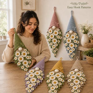 Op de afbeelding: Een collectie van zes gehaakte kabouter-wandhangers in diverse kleuren zoals groen, roze, blauw, paars, geel en beige. Elke kabouter heeft een puntmuts, een ronde neus en een lange baard versierd met witte en gele madeliefjesbloemen.